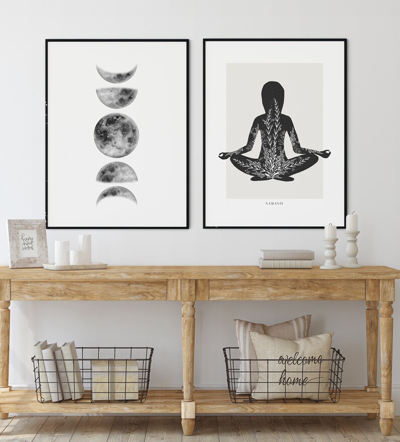 Puede incluir: Dos impresiones enmarcadas en negro en una pared blanca sobre una mesa consola de madera. La impresi&oacute;n de la izquierda muestra una ilustraci&oacute;n en blanco y negro de la luna en diferentes fases. La impresi&oacute;n de la derecha muestra una silueta negra de una persona en una postura de yoga con un dise&ntilde;o floral dentro de la silueta. El texto "SAMASITI" est&aacute; debajo de la imagen. La mesa consola tiene dos cestas de alambre negro en el estante inferior. La cesta de la izquierda contiene libros y la cesta de la derecha contiene una almohada con el texto "welcome home".