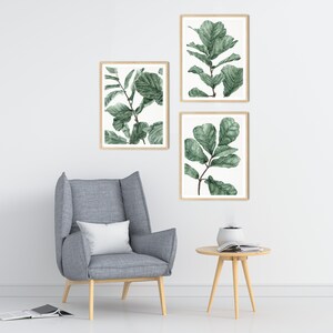 Op de afbeelding: Drie ingelijste aquarelprints van groene bladeren op een witte achtergrond. De prints zijn verticaal in een rij op een witte muur gerangschikt. Een grijze fauteuil met een wit kussen staat voor de prints. Een kleine houten tafel met een tijdschrift en een diffuser staat voor de stoel.