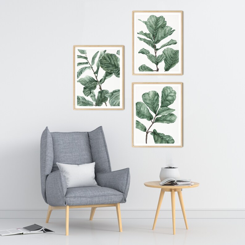 Op de afbeelding: Drie ingelijste aquarelprints van groene bladeren op een witte achtergrond. De prints zijn verticaal in een rij op een witte muur gerangschikt. Een grijze fauteuil met een wit kussen staat voor de prints. Een kleine houten tafel met een tijdschrift en een diffuser staat voor de stoel.