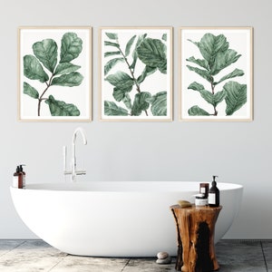 Op de afbeelding: Drie ingelijste aquarelprints van groene bladeren op een witte achtergrond. De prints hangen aan een witte muur boven een wit bad en een houten kruk.
