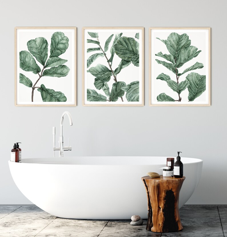 Op de afbeelding: Drie ingelijste aquarelprints van groene bladeren op een witte achtergrond. De prints hangen aan een witte muur boven een wit bad en een houten kruk.