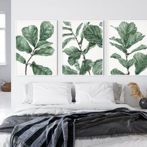 Op de afbeelding: Drie ingelijste aquarelprints van groene bladeren op een witte achtergrond. De prints hangen aan een witte muur boven een bed met witte beddengoed en een grijze deken.