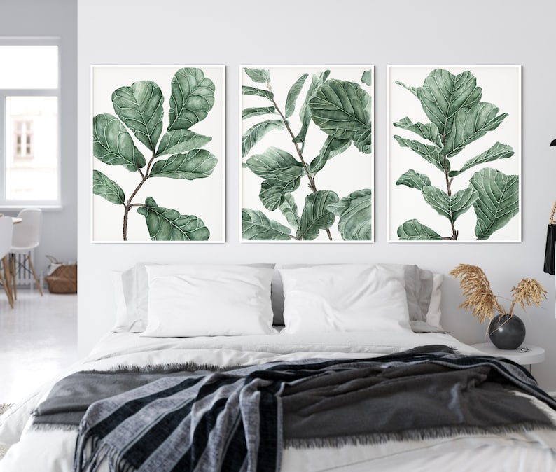 Op de afbeelding: Drie ingelijste aquarelprints van groene bladeren op een witte achtergrond. De prints hangen aan een witte muur boven een bed met witte beddengoed en een grijze deken.