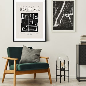 May include: A black and white abstract art print with the text "GALERIE BOHEME" and "Exviva l'arte!" in a white sans-serif font. The print is framed in black and hanging on a white wall. A green velvet armchair with a gray pillow sits in front of the print. A black metal floor lamp with a clear glass globe sits on a black metal stand next to the armchair.