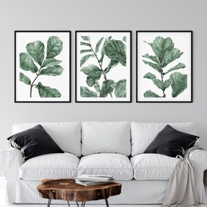 Op de afbeelding: Drie ingelijste aquarelprints van groene bladeren op een witte achtergrond. De prints hangen aan een witte muur boven een witte bank met zwarte kussens.