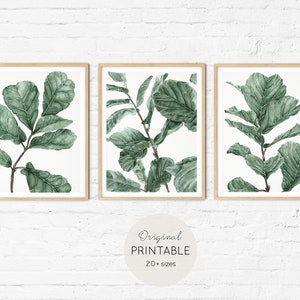 Op de afbeelding: Set van drie printbare kunstprints met aquarel illustraties van groene bladeren. De prints zijn ingelijst in natuurlijke houten frames en worden weergegeven op een witte bakstenen muur.