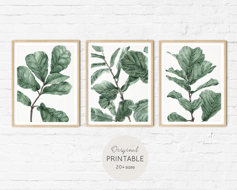 Op de afbeelding: Set van drie printbare kunstprints met aquarel illustraties van groene bladeren. De prints zijn ingelijst in natuurlijke houten frames en worden weergegeven op een witte bakstenen muur.