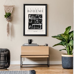 May include: A black and white abstract art print with the text "GALERIE BOHEME" in a black frame hanging on a beige wall above a wooden shelf with a black metal frame. The shelf is holding a black coffee mug and a stack of books. A black wicker stool is in the foreground. A potted plant with green leaves is on the right side of the image.