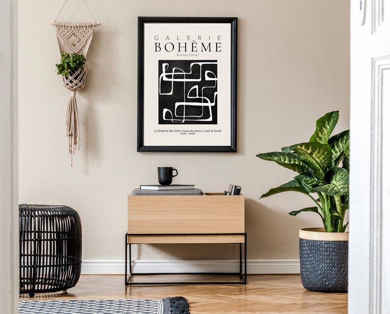 May include: A black and white abstract art print with the text "GALERIE BOHEME" in a black frame hanging on a beige wall above a wooden shelf with a black metal frame. The shelf is holding a black coffee mug and a stack of books. A black wicker stool is in the foreground. A potted plant with green leaves is on the right side of the image.
