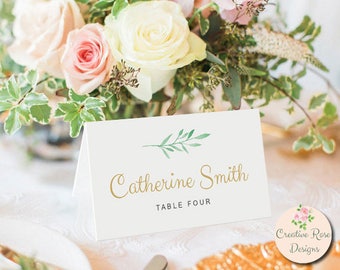 May include: A white place card with gold script that reads "Catherine Smith" and "Table Four". The card has a watercolor green leaf design and is set in front of a floral arrangement with pink and white roses.