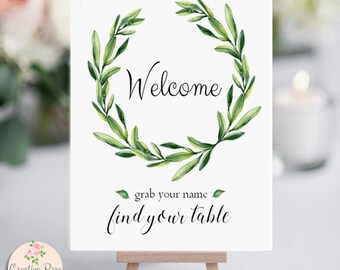 May include: A white sign on a small wooden easel with the word "Welcome" in elegant script, encircled by a watercolor wreath of green leaves. Below, the text reads "grab your name find your table."