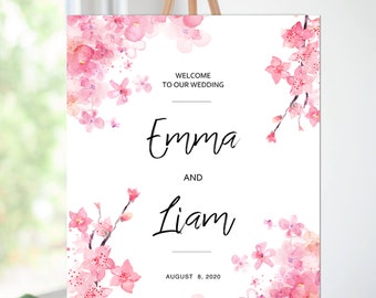 May include: A wedding welcome sign on a wooden easel. The sign is white with pink watercolor cherry blossom accents. The text reads "Welcome to Our Wedding" and the names "Emma and Liam" with the date "August 8, 2020".
