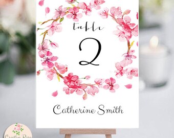 May include: A white table number card with a watercolor floral design. The card features a wreath of pink cherry blossoms around the number "2". The text "Table" and "Catherine Smith" are also displayed. The card is propped up on a small wooden easel.