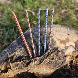 May include: Close-up of five twisted metal rods, some copper-toned and others silver, emerging from a weathered wooden surface. The rods have a spiral design, with the copper ones featuring a reddish-brown hue. The background is blurred, suggesting an outdoor setting.