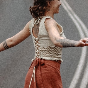 May include: A woman wearing a white crocheted top with a brown and white patterned design and a long, rust-coloured skirt with a geometric pattern. She is standing on a paved road with white lines.