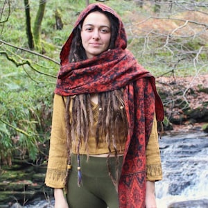 May include: A woman wearing a red and black patterned scarf with a matching hood. She is also wearing a mustard yellow long-sleeved shirt and olive green leggings. The background features a forest and a small waterfall.