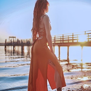 May include: A woman with long red dreadlocks wears a long, rust-colored maxi dress with a side slit and a lace-up back. She stands on a pebbled beach with a wooden pier in the background.