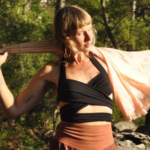 May include: A woman wearing a black wrap top, brown pants, and a light pink shawl with lace trim. The woman is holding a light brown parasol. The background is a forest with tall trees and green foliage.