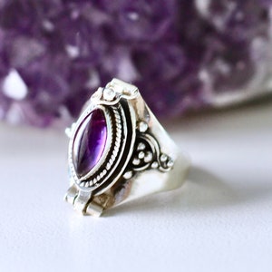 May include: A silver ring featuring a purple amethyst stone set within an intricate filigree design. The ring is positioned on a white surface, with a large amethyst crystal in the background.