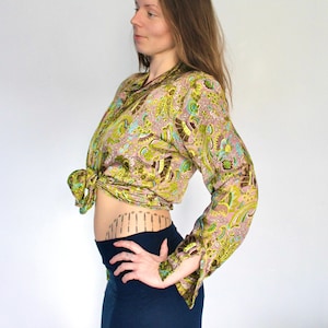 Op de afbeelding: Een vrouw draagt een lang mouwig, groen en bruin paisley print blouse die in de taille is geknoopt. De blouse heeft een lichtblauw en groen bloemenpatroon. Ze draagt een zwarte rok met een zwarte riem.