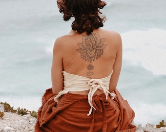May include: A person seen from behind wearing a cream-colored, tie-back crop top and a rust-colored wrap skirt. The person has a large lotus flower tattoo on their upper back and a series of smaller circular chakra symbols along their spine.