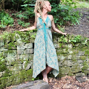 Peut inclure: Une femme portant une longue robe à motif beige et bleu sarcelle avec un décolleté halter. Elle se tient devant un mur de pierre recouvert de mousse et de verdure.