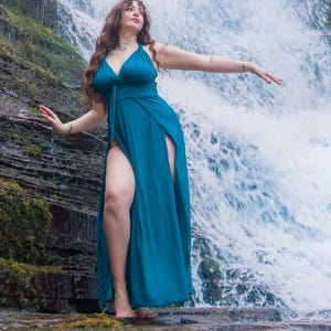 Peut inclure: Une longue robe bleu sarcelle avec un décolleté plongeant et des fentes hautes. La robe est portée près d'une cascade, avec l'eau qui coule en arrière-plan. La personne se tient sur un rocher recouvert de mousse, les bras tendus.