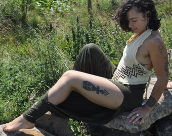 May include: A cream-colored halter top featuring a dark brown geometric zigzag pattern across the midsection, paired with olive green leggings that have a high-cut side slit, worn while sitting on a large stone in a natural outdoor setting.