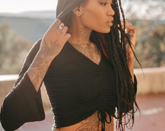 Peut inclure: Une femme avec de longues tresses noires porte un haut noir à capuche avec une taille cintrée et un pantalon jaune moutarde. Elle a plusieurs tatouages sur les bras et le torse.