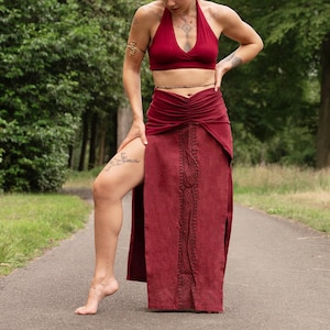 May include: A burgundy halter top and matching skirt. The skirt features a high slit and an embroidered panel down the front. The outfit is made of a soft, flowing fabric, perfect for warm weather. The model is standing on a paved path surrounded by trees.