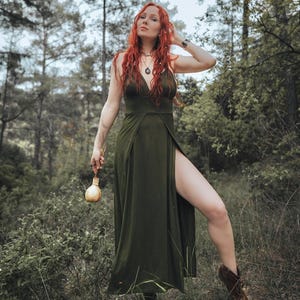 May include: A floor-length olive green dress featuring a deep V-neckline, thin spaghetti straps, and a high side slit that reveals the leg. The garment is worn with dark brown lace-up boots and a small, round, light-colored bottle held in one hand.
