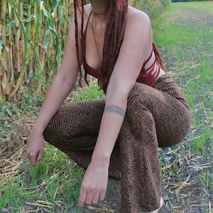 Peut inclure: Une femme avec de longues dreadlocks rouges porte un débardeur rouge et un pantalon à motifs marrons et noirs. Elle est accroupie dans un champ de maïs.