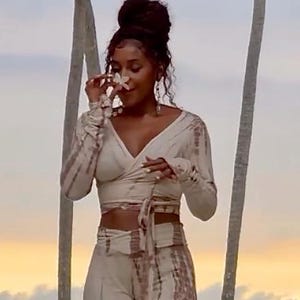 Op de afbeelding: Een tweedelige set bestaande uit een wikkeltop met lange mouwen en een broek met hoge taille en wijde pijpen. Het kledingstuk heeft een lichtbeige basiskleur met een verticaal bruin tie-dye patroon over de gehele stof voor een consistent uiterlijk.