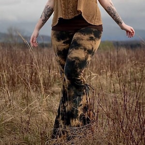 Op de afbeelding: Een vrouw met lang rood haar draagt een bruin gehaakt topje en een zwarte en bruine tie-dye broek met wijde pijpen. Ze staat in een veld met hoog gras.