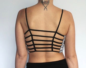 May include: A black crop top featuring a strappy back design with five horizontal black bands connecting the shoulder straps. The sides of the top display a white and black patterned fabric. The garment is worn with a plain black skirt.