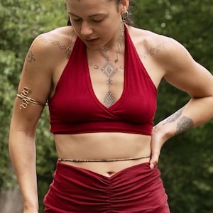 May include: A deep red halter top with a V-neckline paired with a matching deep red skirt featuring a gathered front panel and a central vertical embroidered pattern. The outfit is accessorised with a gold-toned arm cuff and a thin beaded waist chain.