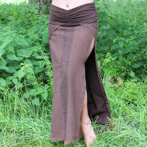 Può includere: Un top avvolgente bordeaux con maniche lunghe e cappuccio, abbinato a una gonna marrone con spacco alto e ricami decorativi. L'outfit è indossato all'aperto in un ambiente naturale.