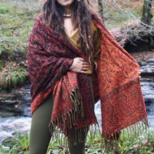 May include: A shawl with a paisley design in shades of red, black, and orange. The shawl has a fringed edge and is draped over the shoulders. The person is wearing a gold-colored top and olive green leggings.