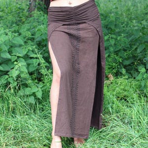 May include: A brown skirt with a high slit and an intricate geometric pattern. The skirt has a ruched waistband and is made of a flowing fabric. The model is standing in a grassy area.
