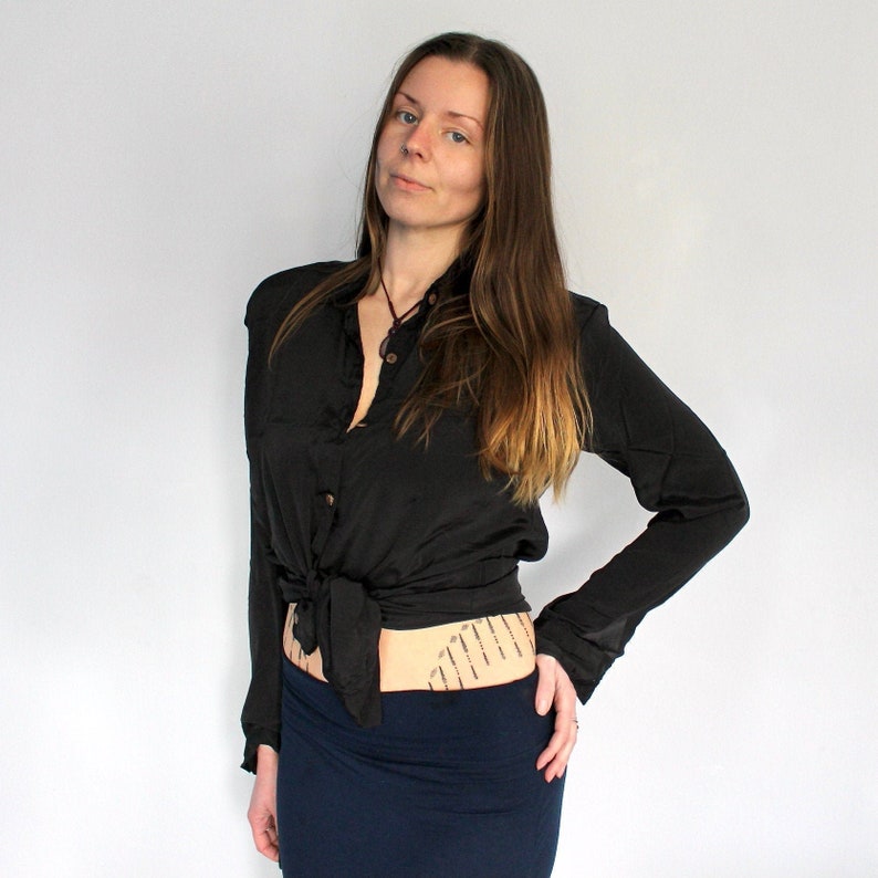 Puede incluir: Una mujer con una camisa negra abotonada atada a la cintura y una falda azul oscuro. Tiene el pelo largo y casta&ntilde;o y mira a la c&aacute;mara.