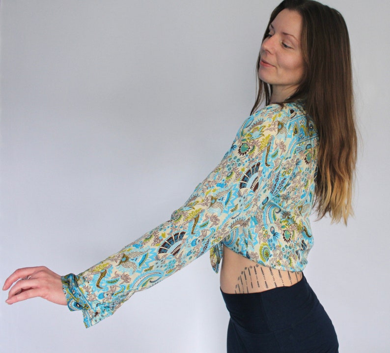 Op de afbeelding: Een vrouw draagt een blauw en wit gebloemd gewashemd met lange mouwen. Het shirt heeft een paisley-ontwerp. De vrouw draagt een donkerblauwe rok.