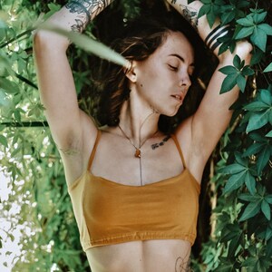 May include: A woman with tattoos on her arms is wearing a mustard yellow crop top and a white skirt. She is standing in front of a wall of green leaves.