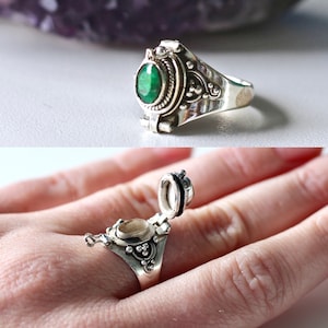 May include: A silver ring with a hinged compartment and a green gemstone. The ring is open, revealing the empty compartment inside.