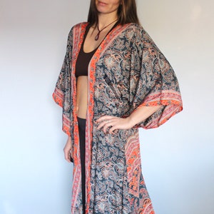 May include: A long, flowing black and orange patterned kimono with a floral design. The kimono has a wide, open neckline and long sleeves. The model is wearing a brown tank top underneath the kimono.