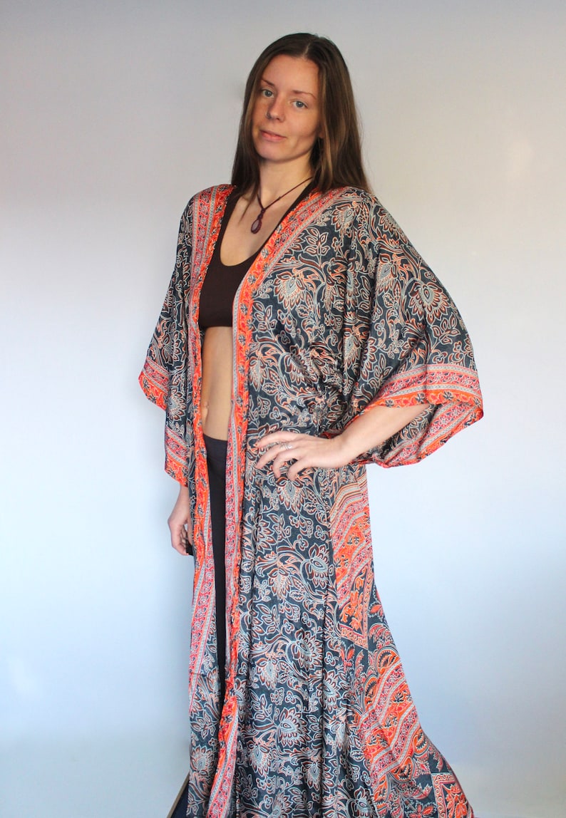May include: A long, flowing black and orange patterned kimono with a floral design. The kimono has a wide, open neckline and long sleeves. The model is wearing a brown tank top underneath the kimono.