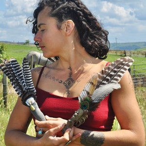May include: A person holding two feather wands. The wands are made of feathers and have a wooden handle. The person is wearing a red top and has a tattoo on their chest.