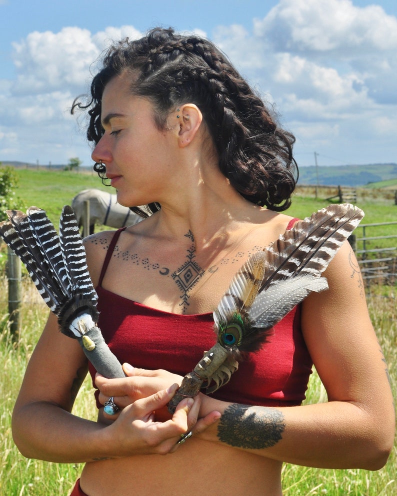 May include: A person holding two feather wands. The wands are made of feathers and have a wooden handle. The person is wearing a red top and has a tattoo on their chest.
