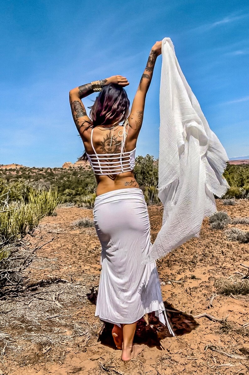May include: A woman wearing a white crop top with a criss-cross back and a white maxi skirt. She is standing in a desert landscape with her arms raised and a white scarf flowing behind her.
