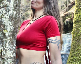 May include: A short-sleeved red crop top with a deep V-neckline and tie-back closure. The garment is made of a solid red fabric and features a fitted silhouette that ends above the waistline, paired with dark purple bottoms.