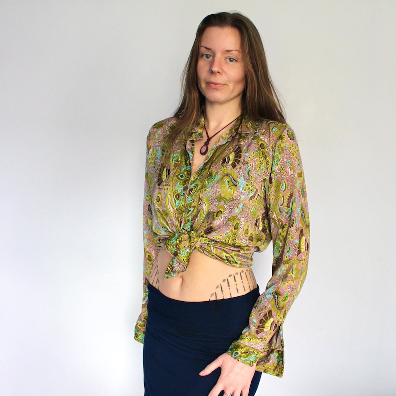 Op de afbeelding: Een vrouw draagt een groen en bruin paisley-print shirt dat in de taille is geknoopt. Het shirt heeft lange mouwen en een kraag. Ze draagt een donkerblauwe rok.